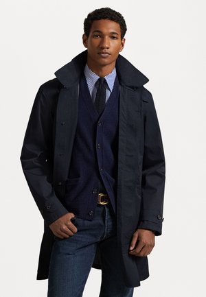 Marineblauwe trenchcoat met grote kraag, knoopsluiting en zijzakken, gedragen over een donkere vest en gestreept shirt. Klassieke spijkerbroek.