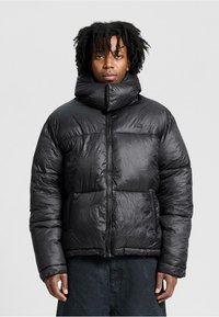 Person iført en skinnende sort pufferjakke med en høj polstret krave, der står foran en ensfarvet lys grå baggrund.