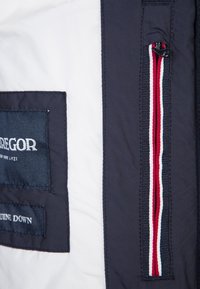 Navy puffer jacket section with white lining, featuring a branded patch and zip pocket with red, white, and blue striped accents.