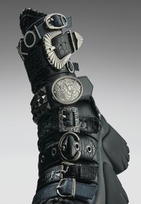 Svarte chunky støvler med flere dekorative sølvspenner, nagler og teksturerte lærreimer mot en grå gradient bakgrunn.