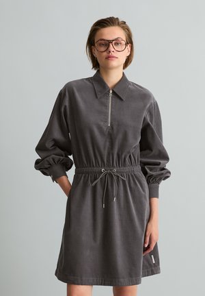 Jong persoon draagt een oversized bruine jurk met ritskraag en trekkoord in de taille, staand tegen een effen lichtgrijze achtergrond.