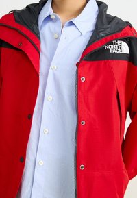 Rote wasserdichte Jacke mit schwarzen Akzenten, mit Reißverschluss, Druckknöpfen und einem Logo auf der Brust. Darunter ein hellblau gestreiftes Hemd.