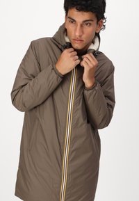 Brown weatherproof jacket with a hood, featuring a plush lining and a front zipper with multicolored stripes. Elastic cuffs and side pockets.