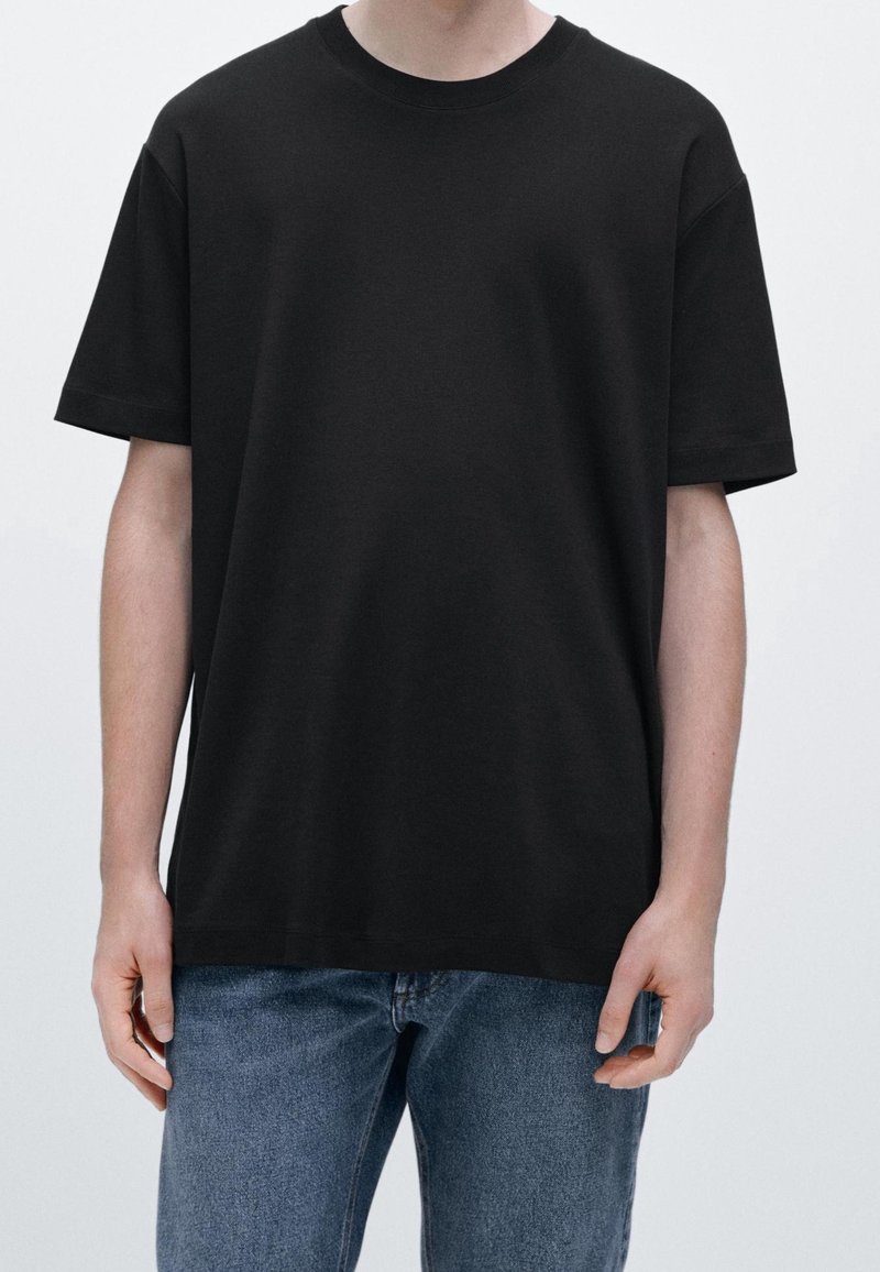 Personne portant un t-shirt noir uni à manches courtes et un jean bleu, debout devant un fond clair uni.