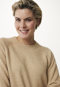 Hellbeige Pullover mit gerippter Textur, rundem Ausschnitt und Raglanärmeln, getragen von einer Person, die vor einem hellen Hintergrund lächelt.