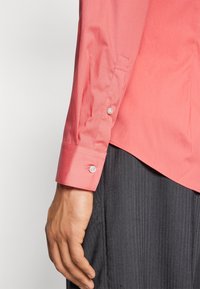 Coral button-up shirt with long sleeves, featuring white buttons and a smooth texture. The shirt has a curved hem and is paired with textured black pants.