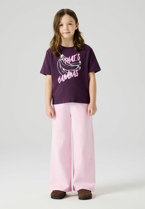 Barn med skulderlangt brunt hår iført en lilla T-shirt med teksten "That's Bananas", lyserøde vide bukser og brune sko, stående på grå baggrund.