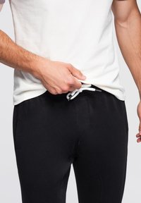Svarta träningsbyxor med dragsko midja, tillverkade av mjukt material, med en enkel design och sidfickor. Modellen justerar midjan.