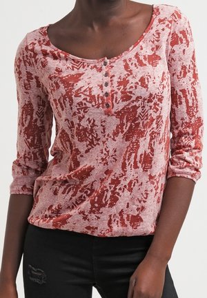 Dames shirt met lange mouwen en ronde hals, voorzien van een rood-wit abstract patroon en vier knopen aan de voorkant, gecombineerd met gescheurde zwarte broek.