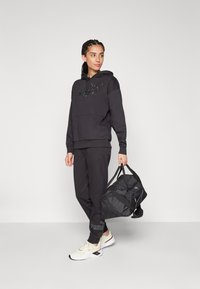 Sudadera negra con logo, pantalones de chándal a juego y zapatillas blancas. El modelo sostiene un bolso de lona negro. Diseño sencillo, ajuste casual, material de mezcla de algodón.