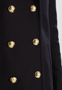 Navy ullkappa med stora guldblickar med präglade mönster, accentuerad med sidofickor och skräddarsydd passform.