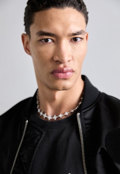 Veste bomber noire avec une fermeture éclair argentée, portée sur un T-shirt noir. Collier ras du cou en perles blanches avec des accents en forme de croix. Texture de peau lisse.