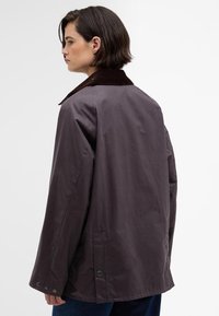 Casaco roxo escuro, ligeiramente oversized, com colarinho de corduroy castanho, botões de pressão e bolsos laterais. Textura suave, design minimalista.