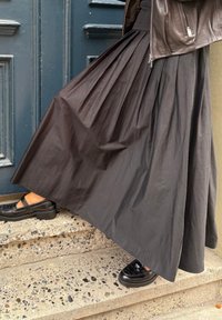 Black pleated maxi skirt made of lightweight fabric, paired with shiny black Mary Jane shoes, displayed against a textured stone staircase.
