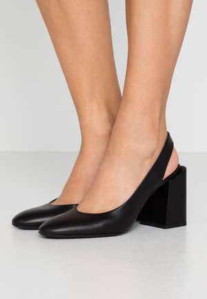 Escarpins slingback en cuir noir avec bouts pointus et talons carrés, portés pieds nus sur un sol et un mur gris clair.