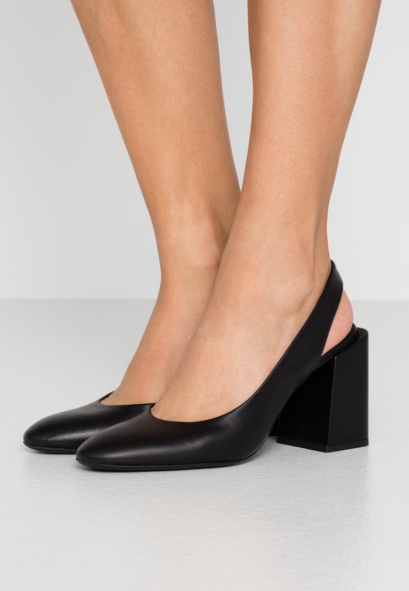 Escarpins slingback en cuir noir avec bouts pointus et talons carrés, portés pieds nus sur un sol et un mur gris clair.