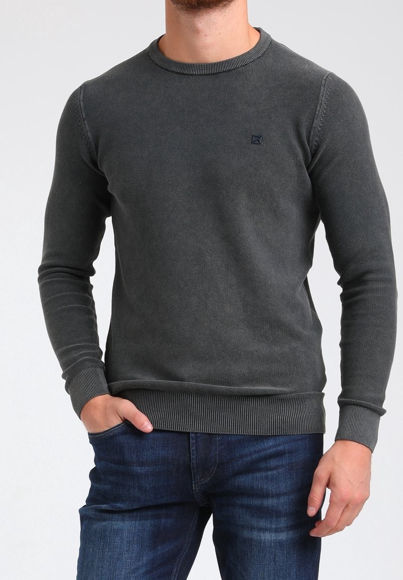 Pull en maille grise avec un col rond et des poignets côtelés, présentant un motif texturé et un petit logo sur la poitrine. Porté avec un jean bleu.