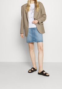 Beige kavaj, vit grafisk t-shirt, denimkjol, svarta sandaler med spänne. Släta tygtexturer, avslappnad design och neutral färgpalett.