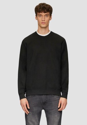 Zwarte gebreide sweatshirt met textuurstof, ronde halslijn en geribbelde manchetten. Draagt over een witte T-shirt. Gecombineerd met grijze versleten jeans.