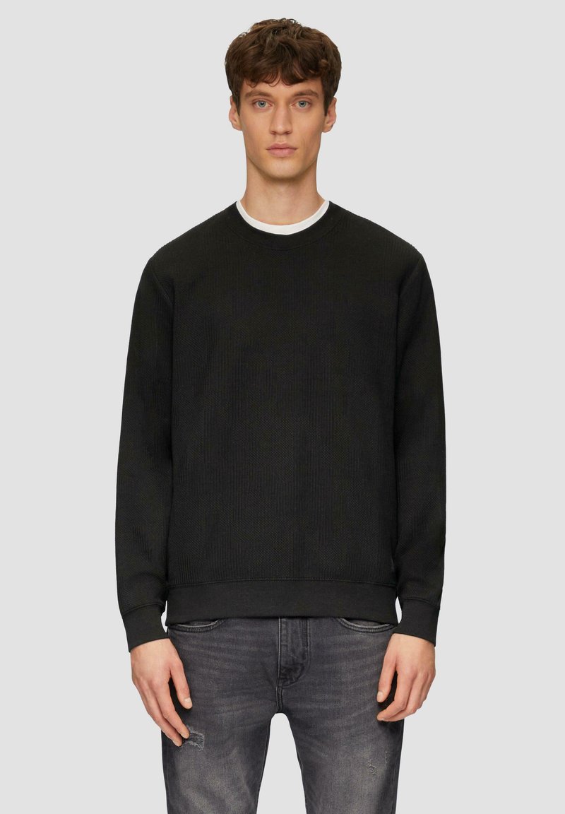 Zwarte gebreide sweatshirt met textuurstof, ronde halslijn en geribbelde manchetten. Draagt over een witte T-shirt. Gecombineerd met grijze versleten jeans.