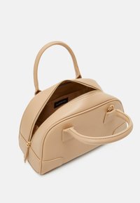 Beige keinokangas käsilaukku, jossa on pyöreä muoto, kaksoiskahvat, vetoketjukiinnitys ja teksturoidut yksityiskohdat sivuilla. Sisäosa on vuorattu.