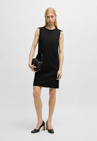 Schwarzes ärmelloses Kleid mit geradem Schnitt und Schulterakzenten. Das Model hält eine kleine schwarze Handtasche und trägt schwarze High Heels.