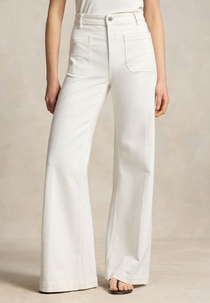 Frau trägt weiße hoch taillierte, weit geschnittene Hose mit aufgesetzten Fronttaschen und Flip-Flop-Sandalen, steht vor neutralem Hintergrund.