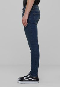 Uske tanke traperice u tamnoplavoj denim boji imaju srednji struk, prednje i stražne džepove te kontrastne šavove. Uparene s crnim tenisicama.