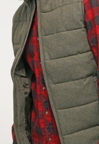 Gepolsterte olivgrüne Weste mit Reißverschluss, die über ein rot-schwarzes kariertes Button-up-Hemd getragen wird und ein glattes Innenfutter aufweist.