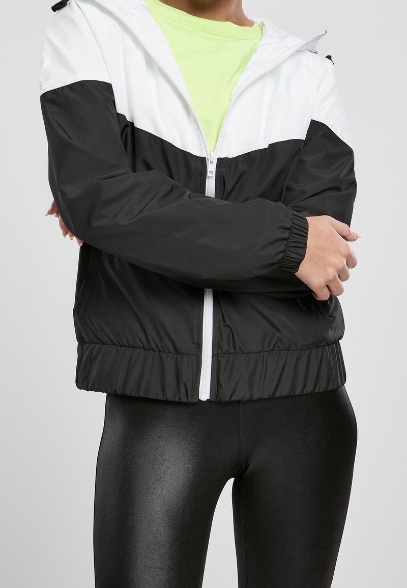 Personne portant une veste zippée noir et blanc sur un t-shirt vert citron avec les bras croisés, associée à des leggings noirs brillants.