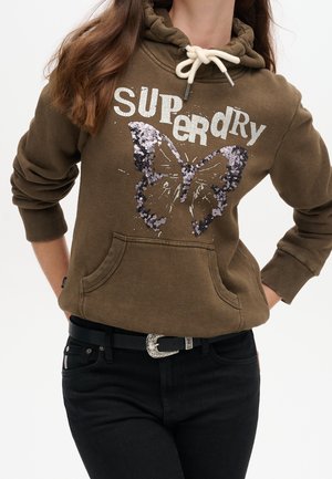 Kobieta w brązowej bluzie z kapturem z napisem "Superdry" i dużym motywem motyla z cekinów, w połączeniu z czarnymi spodniami i paskiem.