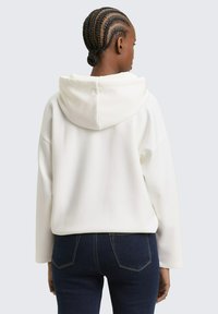 Weißer Hoodie mit lockerem Schnitt, Fronttasche und Kapuze mit Kordelzug, kombiniert mit dunkelblauen Jeans. Glatte Textur mit minimalen Nähten.