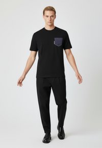 Svart kortärmad t-shirt med en denimficka på vänster bröst, kombinerad med svarta byxor och svarta loafers, stående mot en neutral bakgrund.