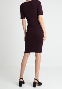 Mörkröd figurnära klänning med rund hals och korta ärmar. Slätt, stretchigt tyg som är designat för att sitta åt kroppen. Bärs med svarta klacksandaler.