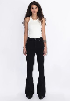 Donna con capelli ricci che indossa un top bianco senza maniche e pantaloni neri svasati, in piedi davanti a uno sfondo bianco, rivolta verso di sé.