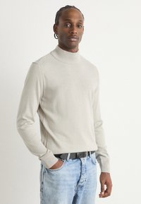 Jersey de cuello alto gris claro en una suave tela de punto, con puños acanalados y un corte casual, combinado con unos vaqueros azul desgastado y un cinturón negro.