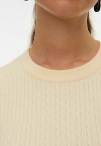 Maglione in cable-knit di un leggero beige, con collo a girocollo a coste e motivo testurizzato. Materiale morbido, design aderente, dettagli minimal.
