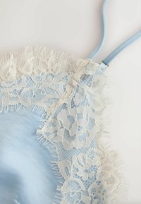 Tissu en satin bleu clair avec une délicate bordure en dentelle florale blanche et une fine bretelle réglable sur un fond uni.