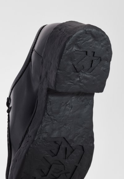 Calzado negro con suela sintética texturizada y un diseño robusto. La parte superior tiene un material suave y detalles de costura visibles.