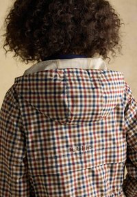 Kind met krullend haar, gekleed in een beige, rood en marineblauw geruite capuchonjas, met de rug naar de camera gekeerd.