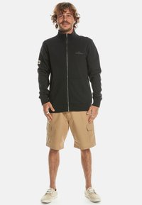 Black zip-up sweatshirt with high collar, combined with beige cargo shorts. Model is wearing light-colored shoes.