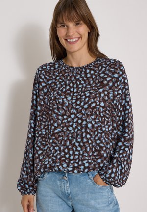 Femme souriante aux cheveux bruns portant un chemisier ample à manches longues à motifs et un jean bleu clair, debout avec une main dans la poche.