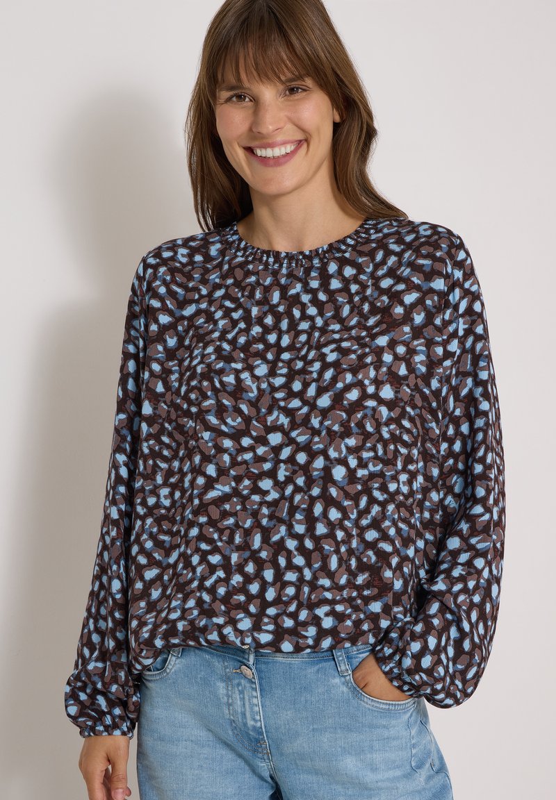 Femme souriante aux cheveux bruns portant un chemisier ample à manches longues à motifs et un jean bleu clair, debout avec une main dans la poche.