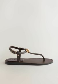 Sandale plate en cuir marron foncé avec bride cheville, fermeture à boucle et breloque dorée en forme de coquillage sur la bride des orteils.