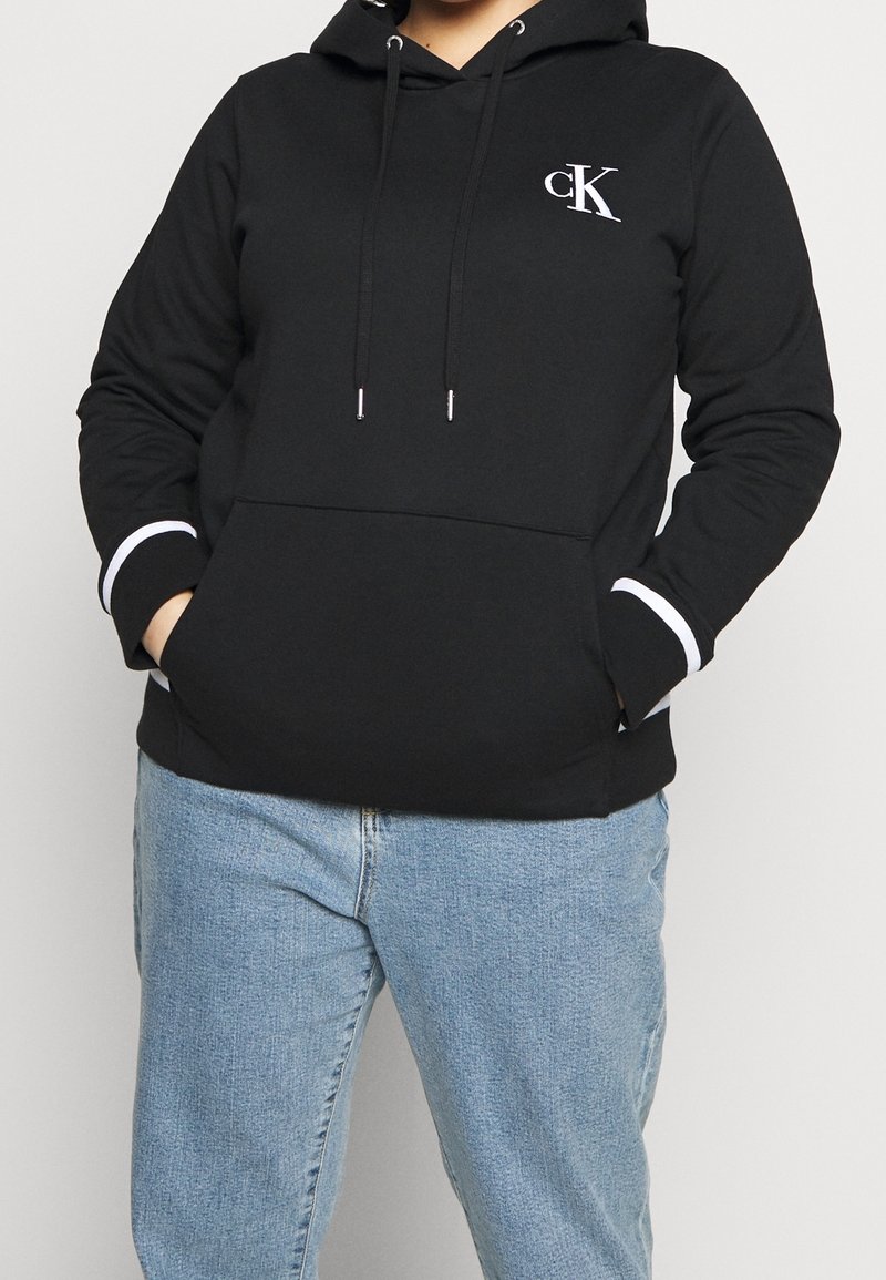Personne portant un sweat à capuche Calvin Klein noir avec un logo blanc sur la poitrine et un jean bleu clair, les mains dans la poche du sweat.