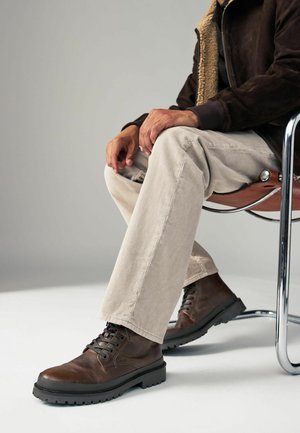 Botas de cuero marrón con cordones y suela de goma, combinadas con pantalones de pana de color claro. El ambiente presenta una silla de cromo y un fondo neutro.