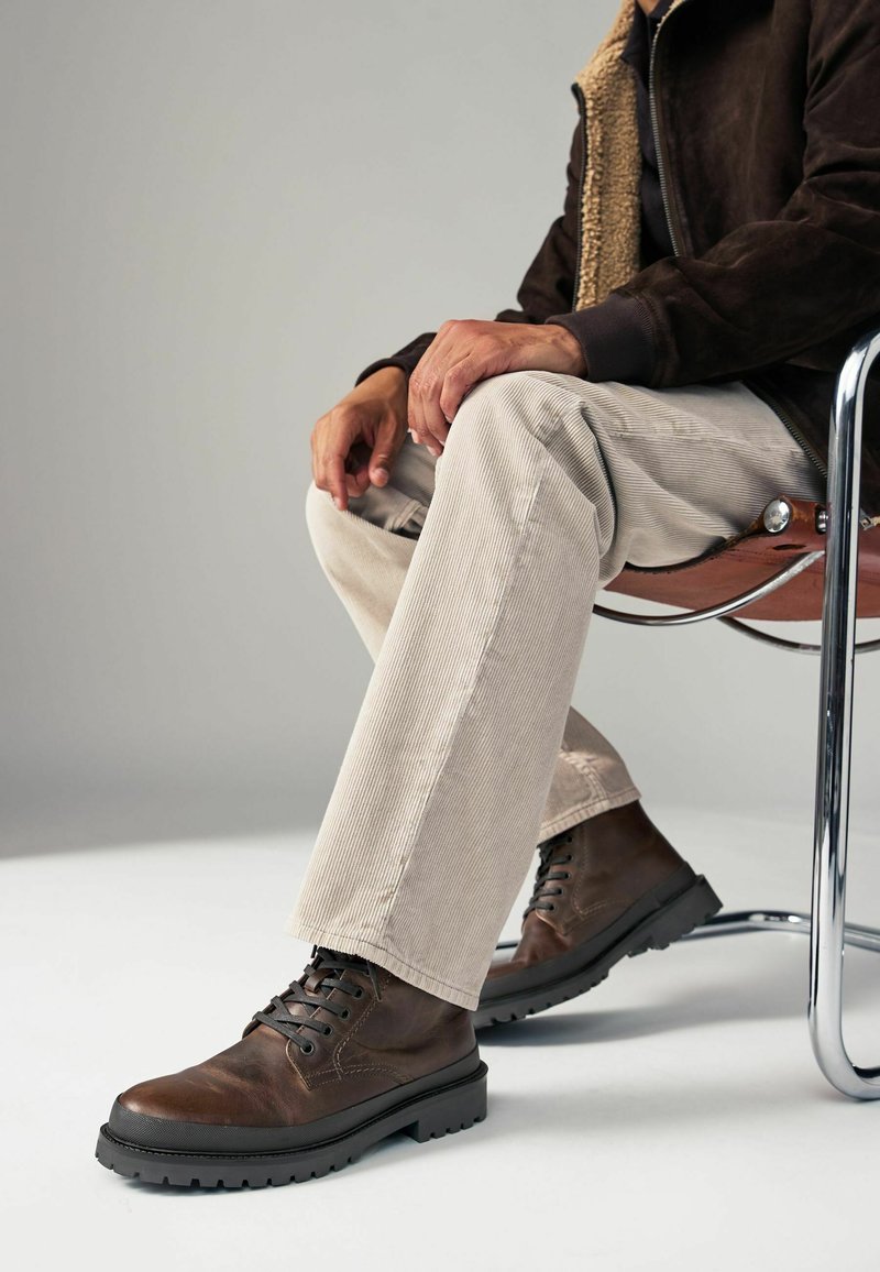 Botas de cuero marrón con cordones y suela de goma, combinadas con pantalones de pana de color claro. El ambiente presenta una silla de cromo y un fondo neutro.