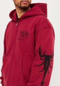 Hombre que lleva una sudadera con cremallera roja y un gráfico de escorpión negro en la manga izquierda, y el texto "Escorpio" en el pecho izquierdo.