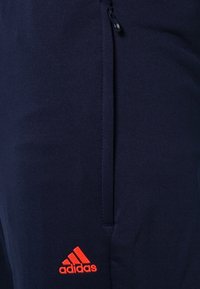 Námořnické sportovní kalhoty s postranním zipem, vyrobené z hladké látky, s jasně červeným logem Adidas blízko lemu.