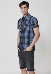 Korte mouwen geruite shirt in blauw en zwart, met twee borstzakken en een knoopsluiting aan de voorkant. Gecombineerd met opgerolde donkergrijze shorts.